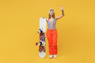 Snowboarder woman wears orange suit goggles mask hat ski costume swimsuit spend extreme weekend waving hand calling isolated on plain yellow background studio. Winter sport hobby trip relax concept.