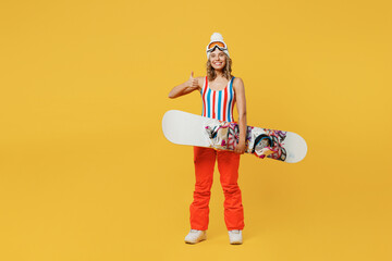 Snowboarder happy fun woman in orange suit goggles mask hat ski costume swimsuit spend extreme weekend show thumb up isolated on plain yellow background studio. Winter sport hobby trip relax concept