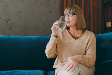 Elderly calm happy fun woman 50s years old wearing casual clothes glasses sits on blue sofa hold glass drink water stay at home flat rest relax spend free spare time in living room indoor grey wall.