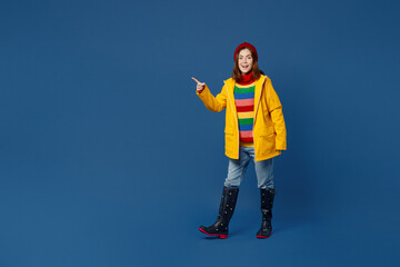 Full body young woman wear sweater red hat yellow waterproof raincoat point index finger aside on...