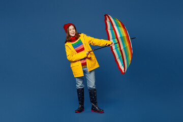 Full body young woman in sweater red hat yellow waterproof raincoat outerwear try to hold opened umbrella isolated on plain dark royal navy blue background Outdoors windy fall weather season concept