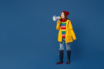 Full body young woman in sweater red hat yellow waterproof raincoat hold scream in megaphone...