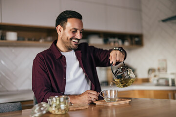 Smiling man sitting at home, pouring a tea into a cup.