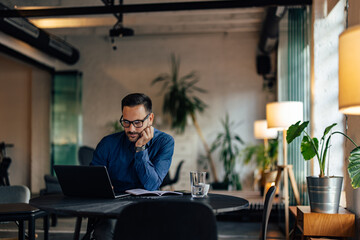 Businessman sitting alone at the office, holding hand on his cheek, using a laptop.