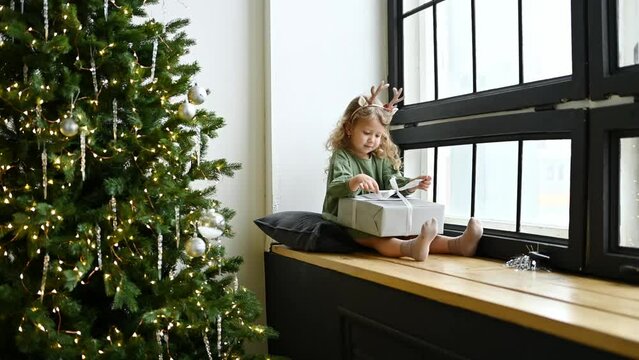 The girl sits on the windowsill by the window and unties the ribbon on the gift