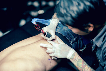 Professional tattooist makes the tattoo on a men waist, focusing on tattoo machines in a modern studio lowlight.