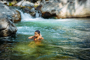 Happy beautiful woman swimming in tropical mountain river with cascade waterfall. Young female traveller bathing in icy water for health improvement and strengthening of immunity.
