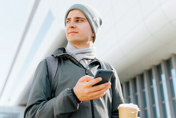 Fototapeta na wymiar Portrait of young man in outerwear and with backpack holding mobile phone and cup of coffee outdoors on sunny day, looking away