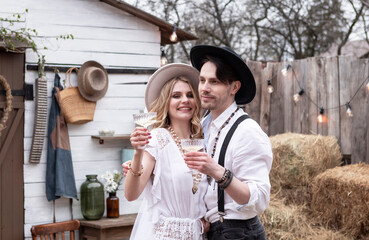 country bride in a white long dress and hat and groom in a white shirt drinking champagne in the village wedding, boho style.