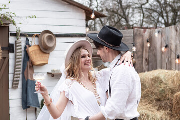 country bride in a white long dress and hat and groom in a white shirt drinking champagne in the village wedding, boho style.