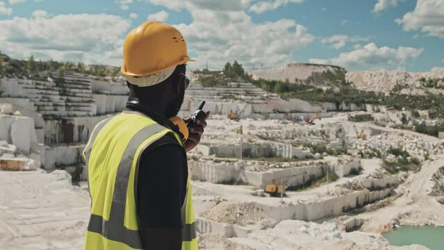Young adult Black man wearing safety helmet, sunglasses working in marble quarry talking to co-worker using handheld transceiver