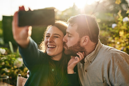 Love, couple and selfie on a patio by happy people kiss, bond and relax at sunset together. Family, phone and man kissing woman for picture while sitting in a backyard, smile and loving at home