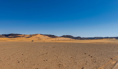 Fototapeta na wymiar Sahara Desert reg stony plain in foreground and sand dune with rocky mountains, 4x4 off-road tiny car far away. Clear blue sky in a sunny day. Tadrart Rouge, Algeria. 