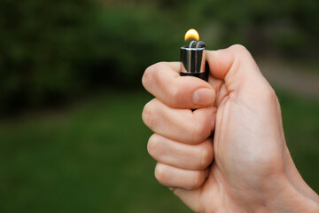 Man holding lighter with burning flame outdoors, closeup. Space for text - obrazy, fototapety, plakaty