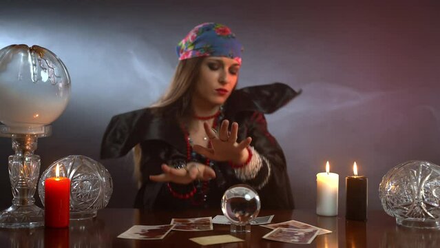 Fiery witch dancing and smiling in her dark illuminated room with mist all over around, local fortune teller sitting at the table with burning candles, round crystal ball and scattered people photos