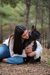 Young caucasian woman hugging her american staffordshire and french bulldog mixed breed dog at a pine forest.