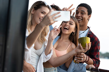 Fototapeta premium Group of multiracial friends on river boat enjoying in summer party, drinking cocktails and taking selfies