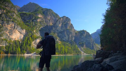 Footage at Lago di Braies, up the mountains in European Alps in Italian Dolomites. Video of a glaciar lake with a man infront, filmed in 4k with a camera on a tripod and nature view in the background
