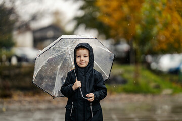 A smiley little boy is standing on the rain in his raincoat and umbrella on a city street at autumn.