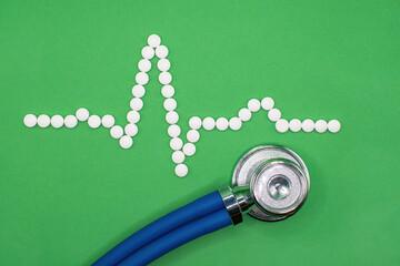 ECG wave form made of medical pills against a green background. A stethoscope sits below the heart...