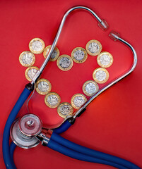 A stethoscope sits on a red background with a heart shape made out of pound coins. Cost of...