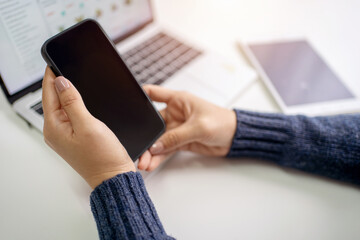Woman is using a smartphone with a blank screen and a laptop on a nearby table. She is sitting in the office wearing a sweater.