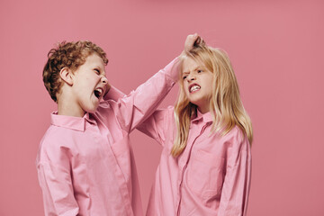 a boy and a girl of school age in pink clothes stand on a pink background and fight fiercely with...