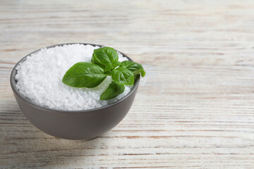 Bowl of natural sea salt and basil leaf on white wooden table, space for text