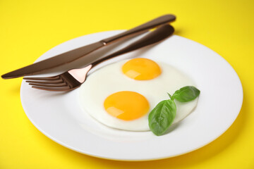 Tasty fried eggs with basil in plate on yellow background, closeup