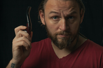 Lumberjack brutal beard muscled man in red shirt with smoking tube pipe standing on dark background