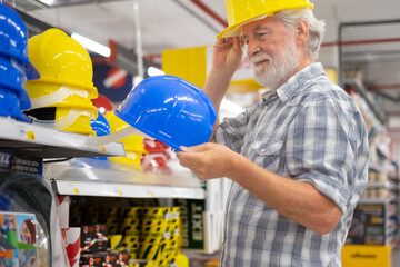 Senior bearded man in hardware store selects and tries a construction helmet. Elderly expert male...