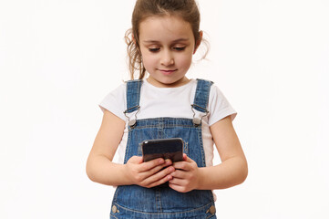 Adorable school age Caucasian little child, cute baby girl wearing blue denim overalls, using mobile phone, browsing website, checking content, isolated over white background with copy space