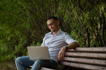 Portrait of a happy entrepreneur in a white shirt and glasses, holding a laptop, sitting on a park bench. Young handsome businessman working remotely in the city