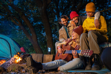Friends at campsite are singing songs, laughing and cooking food on fire while happy smiling girl is playing the guitar. Camping, music, nature and friendship concept. High quality photo