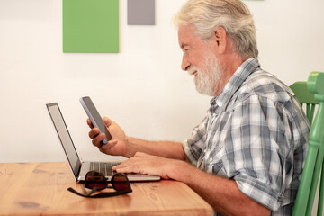 Senior caucasian man sitting at a wooden table in coffee shop using laptop holding his smart phone. Elderly man reading message on cellphone while typing on keyboard