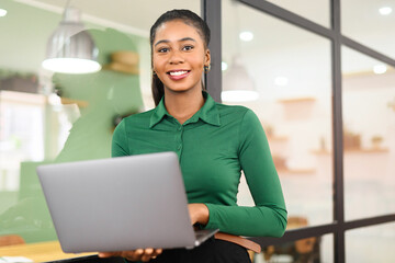 African-American businesswoman in smart casual wear standing in modern coworking space and holding laptop, female freelancer or student with laptop computer indoors smiling cheerfully