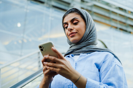 Young muslim woman using cellphone while standing on escalator outdoors