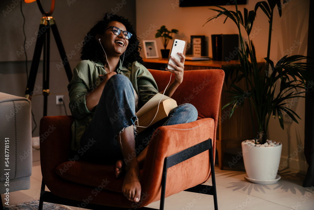 Wall mural African woman in earphones using smartphone while sitting on armchair at home