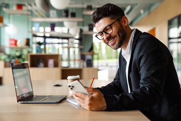 Obraz premium Young businessman using laptop and mobile phone while sitting in office