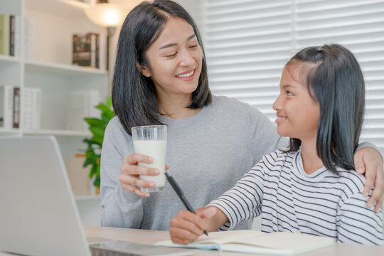 Homeschool Asian young little girl learn, read and do homework with kind mother help, teach and encourage. Mom pass on a glass of milk to daughter. Girl happy to study and education together with mom