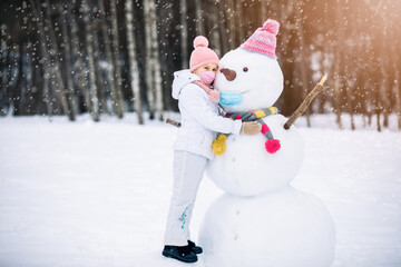 A child girl wearing a mask herself and on a snowman ,playing with a snowman on a winter walk in...