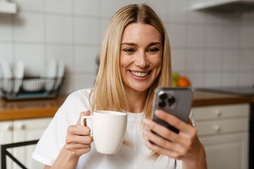 Blonde white woman drinking coffee and using cellphone in kitchen