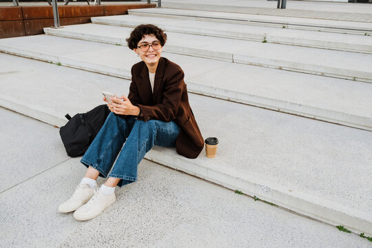 European woman using cellphone and smiling while sitting on stairs