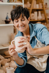 Mature white potter woman holding ceramic jug at the workshop indoors