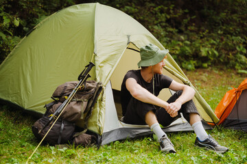 White young man resting in tent while hiking in green forest - obrazy, fototapety, plakaty