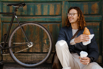 Ginger man holding croissant and cup of coffee while sitting on city street