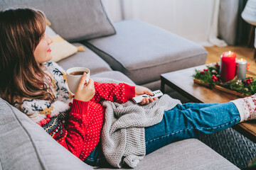Relaxed woman in plaid and warm socks drinking hot tea with remote controller on sofa at home while watching movie, TV with christmas decoration atmosphere. Cozy and comfortable winter concept.