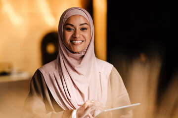 Arabian woman smiling and holding tablet computer while standing indoors