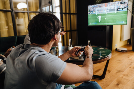 Back View Of Young White Man Playing Football Video Game With Gamepad