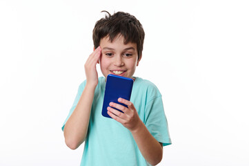 Happy excited teenage school boy holding his hand on his temple, expressing surprise while reading news on his smartphone, isolated over white background with copy space for text